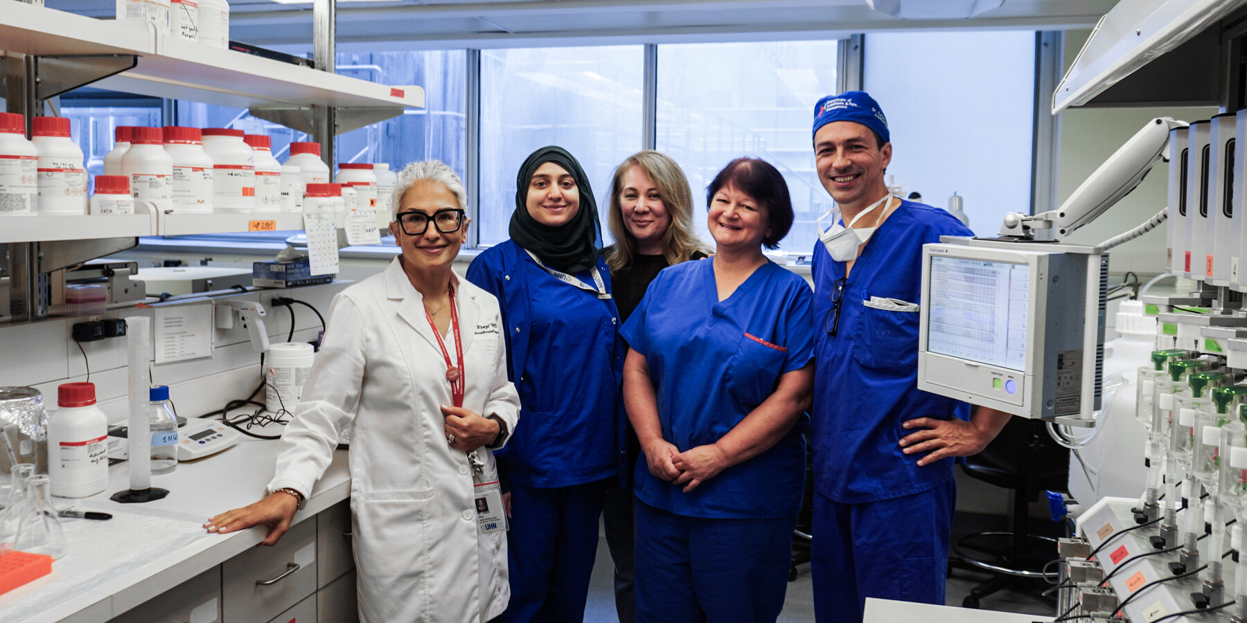 Five people stand in a lab, all wearing scrubs, smiling at the camera