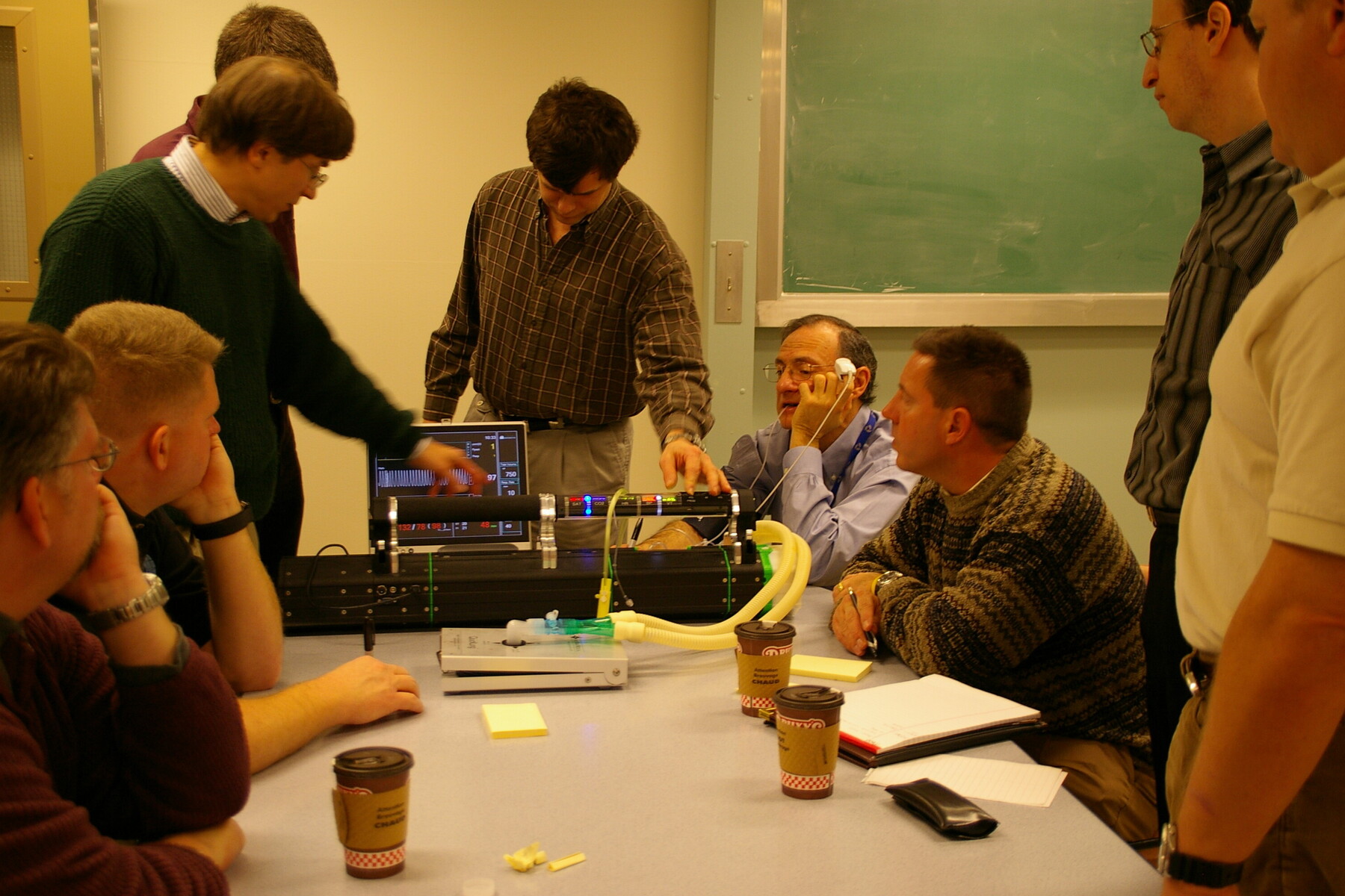 A group of researchers around a table, with one researcher testing the machine. The tester is wearing a finger sensor.